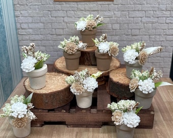 May include: Miniature potted flower arrangements with white, beige, and green floral accents. The small pots are in shades of brown and beige, arranged on wooden platforms and a small wooden pallet. A framed print of a wave is in the background.