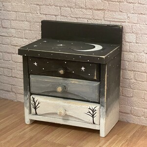 May include: A small wooden nightstand with three drawers. The top drawer is painted black with white stars and a crescent moon. The middle drawer is painted gray and the bottom drawer is painted white with black trees. The nightstand is distressed and has a rustic look.