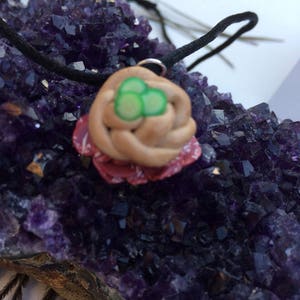 May include: A brown and pink clay bird's nest pendant with three green eggs, hanging on a black cord. The pendant is resting on a bed of purple amethyst crystals.