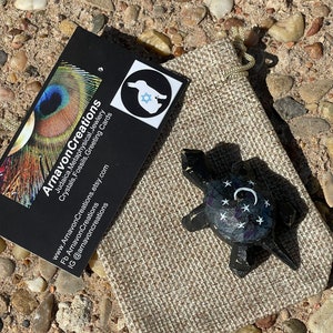 K&ouml;nnte beinhalten: Eine kleine, schwarze Schildkr&ouml;tenfigur mit wei&szlig;en Sternen und einem Halbmond, die auf ihren Panzer gemalt sind. Die Schildkr&ouml;te sitzt in einem kleinen braunen Jutesack.