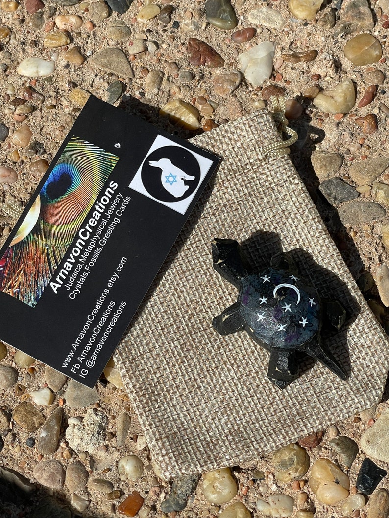 K&ouml;nnte beinhalten: Eine kleine, schwarze Schildkr&ouml;tenfigur mit wei&szlig;en Sternen und einem Halbmond, die auf ihren Panzer gemalt sind. Die Schildkr&ouml;te sitzt in einem kleinen braunen Jutesack.