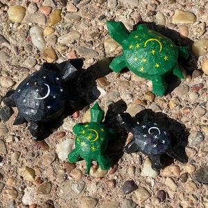 K&ouml;nnte beinhalten: Vier h&ouml;lzerne Schildkr&ouml;tenfiguren mit bemalten Designs. Zwei Schildkr&ouml;ten sind gr&uuml;n mit gelben Sternen und einem Halbmond. Zwei Schildkr&ouml;ten sind schwarz mit wei&szlig;en Sternen und einem Halbmond.