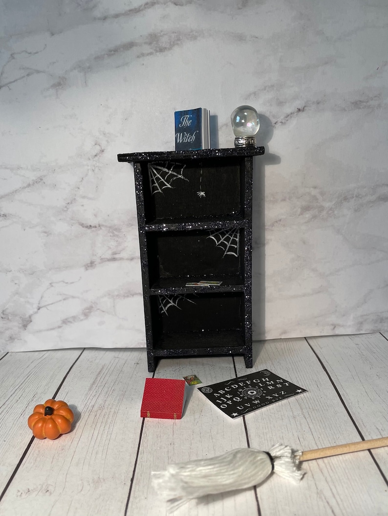 May include: A black bookshelf with three shelves decorated with glitter and spiderwebs. The top shelf has a book titled "The Witch" and a crystal ball. A red book, a pumpkin, a white broom, and a black and white Ouija board are on the floor in front of the bookshelf.