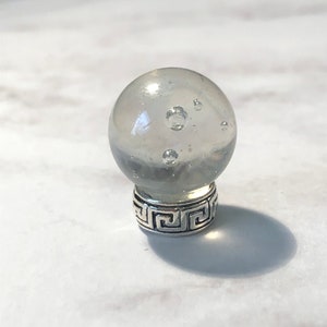 May include: A clear glass sphere with air bubbles inside, mounted on a silver-colored base with a Greek key design. The sphere is partially translucent, and the base has a decorative pattern. The object casts a shadow.