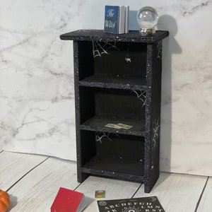 May include: A black wooden bookshelf with three shelves decorated with spiderwebs and glitter. The top shelf has a book titled "The Witch" and a glass crystal ball. The bottom shelf has a small red book and a black and white Ouija board with the letters "ABCDEFGHIJKLMNOPQRSTUVWXYZ" and the words "GOOD BYE".