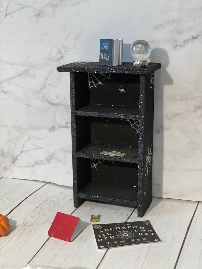 May include: A black wooden bookshelf with three shelves decorated with spiderwebs and glitter. The top shelf has a book titled "The Witch" and a glass crystal ball. The bottom shelf has a small red book and a black and white Ouija board with the letters "ABCDEFGHIJKLMNOPQRSTUVWXYZ" and the words "GOOD BYE".