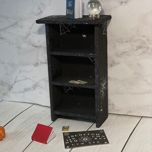 May include: A black wooden bookshelf with three shelves decorated with white spiderwebs and glitter. The top shelf has a book titled "The Witch" and a glass globe. The bottom shelf has a small red book and a black and white Ouija board with the letters A-Z, numbers 0-9, and the words "GOOD BYE".