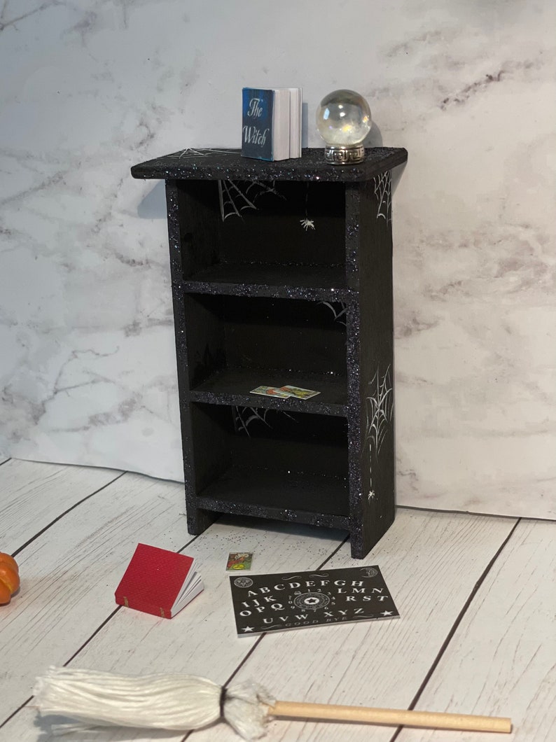 May include: A black wooden bookshelf with three shelves decorated with white spiderwebs and glitter. The top shelf has a book titled "The Witch" and a glass globe. The bottom shelf has a small red book and a black and white Ouija board with the letters A-Z, numbers 0-9, and the words "GOOD BYE".