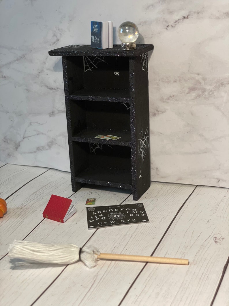 May include: A black wooden bookshelf with three shelves decorated with white spiderwebs and glitter. A small book titled "The Witch" and a crystal ball are on the top shelf. A red book, a green and yellow book, and a Ouija board are on the floor in front of the bookshelf. A white broom is lying on the floor.
