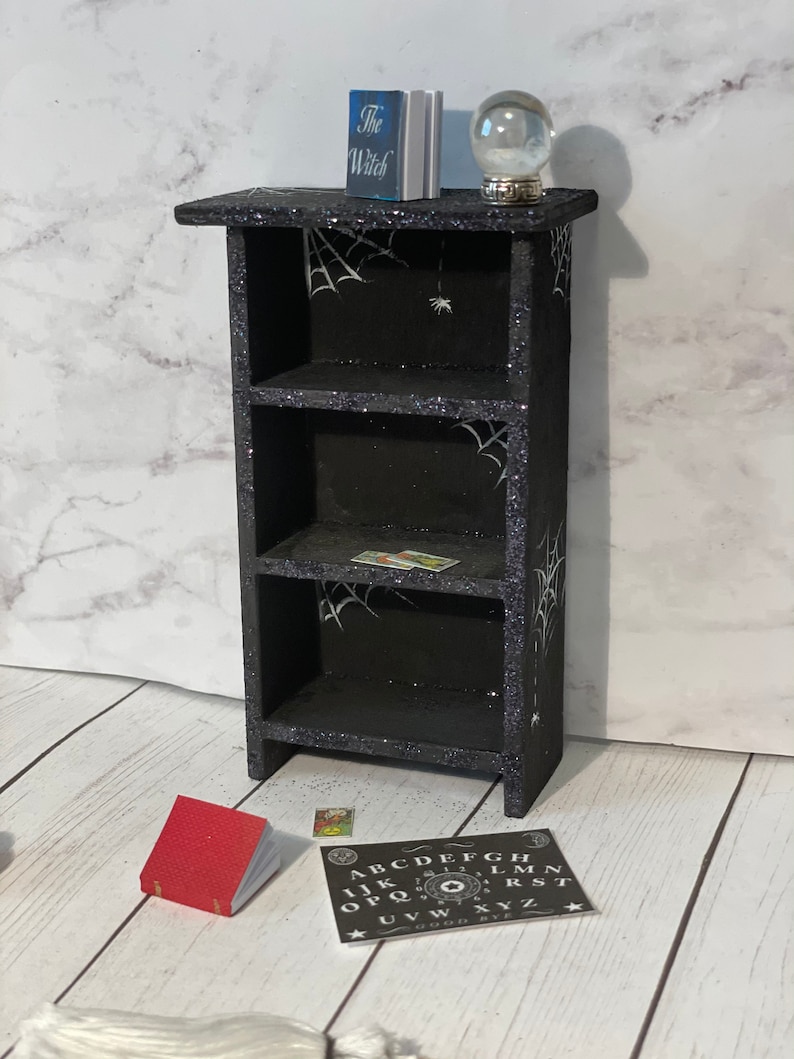 May include: A black wooden bookshelf with three shelves, decorated with white spiderwebs and glitter. The top shelf has a book titled "The Witch" and a glass globe. The bottom shelf has a red book and a small black and white Ouija board.