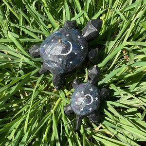 K&ouml;nnte beinhalten: Zwei h&ouml;lzerne Schildkr&ouml;tenfiguren mit einem schwarzen und wei&szlig;en Nachthimmel-Design. Die gr&ouml;&szlig;ere Schildkr&ouml;te hat einen Halbmond und Sterne auf ihrem Panzer bemalt. Die kleinere Schildkr&ouml;te ist ein &auml;hnliches Design, aber kleiner.