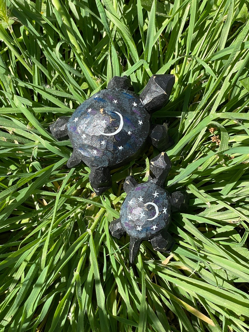 K&ouml;nnte beinhalten: Zwei h&ouml;lzerne Schildkr&ouml;tenfiguren mit einem schwarzen und wei&szlig;en Nachthimmel-Design. Die gr&ouml;&szlig;ere Schildkr&ouml;te hat einen Halbmond und Sterne auf ihrem Panzer bemalt. Die kleinere Schildkr&ouml;te ist ein &auml;hnliches Design, aber kleiner.