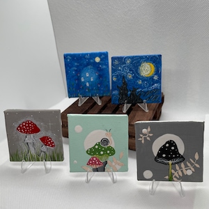 May include: Five small canvas paintings on easels, each depicting a different scene. One painting features a starry night sky with a crescent moon, another shows a mushroom with a snail on top, and another has a mushroom with white dots. The remaining two paintings feature mushrooms with red dots.