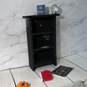 May include: A black bookshelf with three shelves decorated with spiderwebs and a small spider. The bookshelf is sitting on a white wood floor. There is a small red book, a pumpkin, a crystal ball, and a Ouija board on the floor in front of the bookshelf. The bookshelf has a sign on top that says "The Witch".