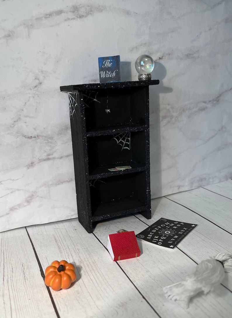 May include: A black bookshelf with three shelves decorated with spiderwebs and a small spider. The bookshelf is sitting on a white wood floor. There is a small red book, a pumpkin, a crystal ball, and a Ouija board on the floor in front of the bookshelf. The bookshelf has a sign on top that says "The Witch".