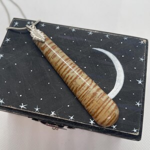 May include: A long, thin, brown and white striped pendant with a silver leaf design hangs from a silver chain. The pendant is resting on a black box with a white crescent moon and stars painted on it.