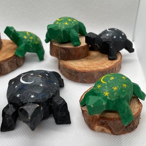 K&ouml;nnte beinhalten: F&uuml;nf h&ouml;lzerne Schildkr&ouml;tenfiguren mit bemalten Designs. Jede Schildkr&ouml;te hat ein anderes Design mit einer Sichelmond und Sternen. Zwei Schildkr&ouml;ten sind gr&uuml;n und drei sind schwarz bemalt.