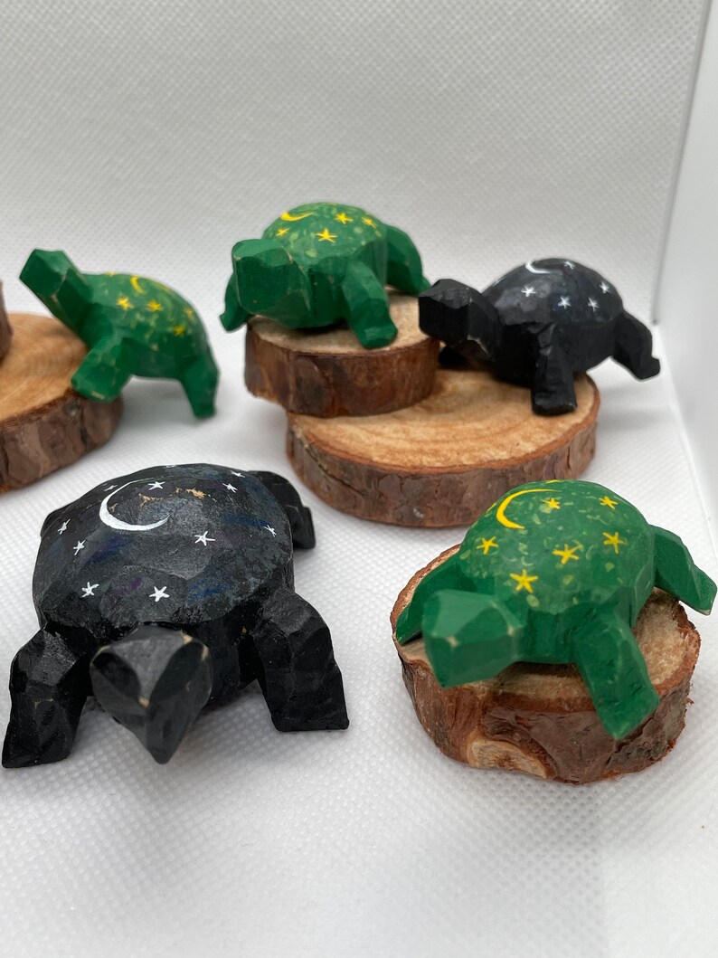 K&ouml;nnte beinhalten: F&uuml;nf h&ouml;lzerne Schildkr&ouml;tenfiguren mit bemalten Designs. Jede Schildkr&ouml;te hat ein anderes Design mit einer Sichelmond und Sternen. Zwei Schildkr&ouml;ten sind gr&uuml;n und drei sind schwarz bemalt.