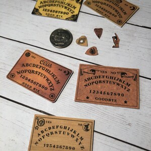 May include: Five miniature Ouija boards with a brown background and black lettering. The boards have the letters A through Z, numbers 0 through 9, and the words "YES", "NO", "GOODBYE", and "OUIJA". There are also four small wooden planchette pieces with different designs.