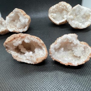 May include: Six open geodes with a rough, tan exterior and a sparkling, white crystal interior are arranged on a dark gray surface. The geodes vary in size and shape, showcasing natural formations.