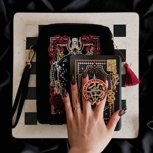 Op de afbeelding: Zwarte fluwelen clutch met rode en gouden borduursels, een zwarte etui met een gouden sluiting en een rode en gouden slangenring. De clutch heeft een gouden polsband en ligt op een zwart-wit geblokt oppervlak.