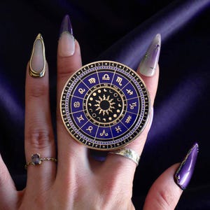 May include: A circular, gold-coloured zodiac wheel with a blue centre, displaying astrological symbols and a sunburst design. The wheel is held by a hand with long, purple and gold-accented nails. The background is a dark, textured fabric.