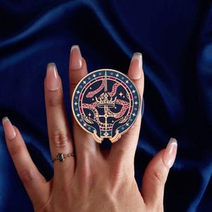 May include: A circular phone grip in navy blue with a gold trim and an intricate design. The design includes a crown, snakes, and hands, with pink and gold detailing. The grip is held by a hand, with a ring visible.