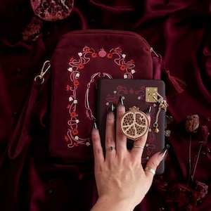 Puede incluir: Bolso de mano y funda de teléfono de terciopelo burdeos con bordado floral. Un charm en forma de granada en tono dorado está unido a la funda del teléfono. El bolso de mano tiene una cremallera dorada y una correa de muñeca.