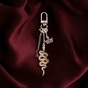 Op de afbeelding: Een goudkleurige sleutelhanger met een slangbedel, een kleine kroonbedel en een rode edelsteen. De sleutelhanger heeft een karabijnhaak en een kettingdetail. De achtergrond is een diep bordeauxrode stof.
