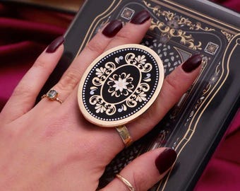 Puede incluir: Un soporte para teléfono de forma ovalada con fondo negro y borde dorado, con un diseño floral blanco. El soporte se sostiene en un dedo, mostrando su patrón decorativo. El fondo incluye un libro oscuro con detalles dorados.