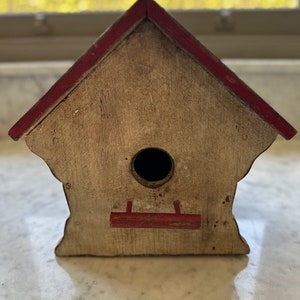 Puede incluir: Una casita de pájaros de madera con un techo rojo y una pequeña abertura para que los pájaros puedan entrar. La casita tiene una percha para que los pájaros puedan posarse.