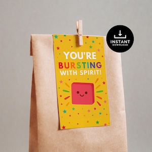 Op de afbeelding: Een gele cadeaulabel met de tekst "YOU'RE BURSTING WITH SPIRIT!" en een lachend rood vierkant. Het label is versierd met kleurrijke sterren en is met een houten clip aan een bruine papieren zak bevestigd.