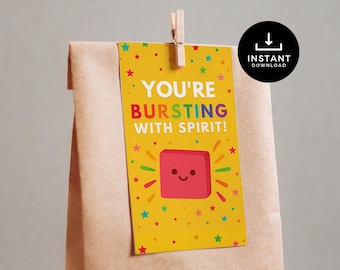 Może przedstawiać: Żółta etykieta prezentowa z napisem "YOU'RE BURSTING WITH SPIRIT!" i uśmiechniętym czerwonym kwadratem. Etykieta jest ozdobiona kolorowymi gwiazdkami i przypięta do brązowej papierowej torby drewnianym klipsem.