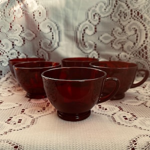 Könnte beinhalten: Ein Set aus fünf rubinroten Teetassen aus Glas. Jede Tasse hat eine abgerundete Schale, einen kleinen Henkel und einen Sockel. Die Tassen stehen auf einer weißen Spitzendecke.