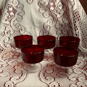 Può includere: Set di cinque coppette da dessert in vetro rosso rubino. Ogni coppetta ha una forma rotonda e bassa e un gambo e una base in vetro trasparente. Sono disposte su una tovaglia di pizzo bianco con sfondo rosso.