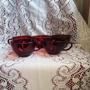 Puede incluir: Un juego de cuatro tazas de té de vidrio rojo rubí con asas. Las tazas están dispuestas sobre un mantel de encaje blanco con un patrón floral. Las tazas son de un color rojo intenso y el encaje añade un contraste delicado.