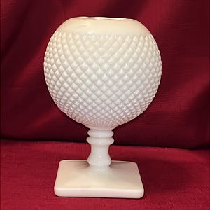 Może przedstawiać: Biały, szklany wazon w stylu vintage z kulistym wierzchołkiem o rombowym wzorze. Kula spoczywa na podstawie cokołu z kwadratowym dnem. Wazon wykonany jest z nieprzezroczystego szkła i umieszczony na czerwonym tle.