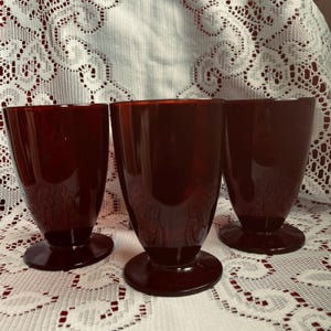 May include: Three ruby red vintage drinking glasses. Each glass has a rounded body and a circular base. The glasses are made of a translucent material, and the light shines through them. The glasses are arranged in a row on a white lace tablecloth.
