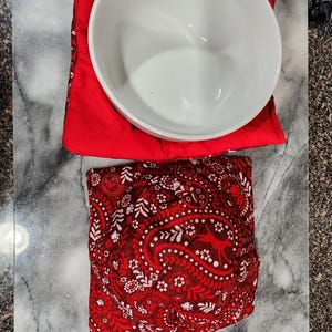 Puede incluir: Un cuenco de cerámica blanco descansa sobre un salvabols rojo. Debajo, un segundo salvabols rojo con un estampado de cachemira es visible. Los artículos están sobre una superficie con vetas de mármol.