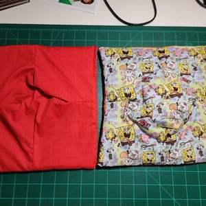 Könnte beinhalten: Zwei quadratische Stoffartikel. Einer ist einfarbig rot. Der andere ist mit SpongeBob Schwammkopf-Figuren auf hellblauem Hintergrund gemustert. Die Artikel liegen auf einer grünen Schneidematte.