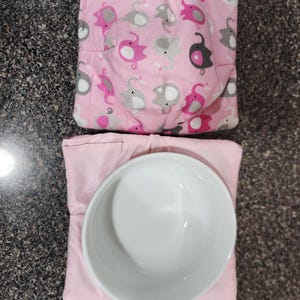 May include: Two bowl cozies. The top one is pink with a pattern of grey and pink elephants. The bottom one is plain pink, with a white bowl inside. The cozies are designed to hold bowls.