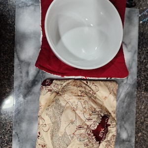 May include: A white ceramic bowl sits atop a red, quilted bowl cozy. Below, a second bowl cozy features a map design in tan and burgundy. The items rest on a gray and white marble cutting board.