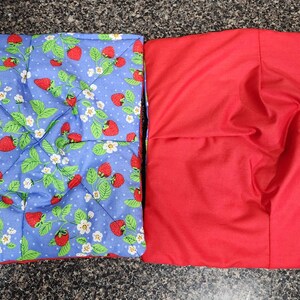 Pode incluir: Duas proteções quadradas para tigelas. Uma é azul com um padrão de morangos e bolinhas brancas. A outra é vermelha lisa. A proteção de morango tem morangos vermelhos, folhas verdes e flores brancas.