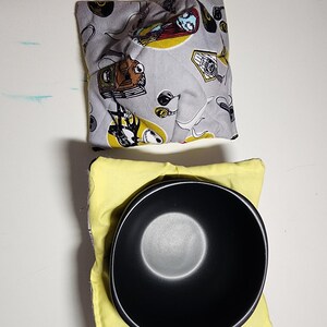 Puede incluir: Un cuenco negro descansa sobre un salvamanteles de tela amarilla. Arriba, un salvamanteles gris presenta un estampado con personajes de dibujos animados e instrumentos musicales. Los salvamanteles están diseñados para proteger las manos de los cuencos calientes.