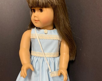 May include: A light blue dress with white lace trim and a matching light blue purse. The dress has a scoop neck and a sleeveless design. The doll is wearing the dress and has brown hair.