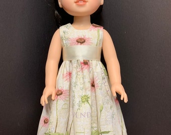 May include: A doll wearing a sleeveless dress with a floral print and a white sash. The dress has a light green background with pink flowers and green leaves. The doll has black hair and brown eyes.