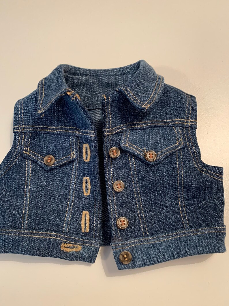 May include: A blue denim vest with a collared neckline and multiple button closures. The vest has two front pockets and a single button on the bottom.