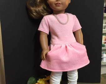 May include: A pink dress with a pocket and white leggings on a doll with brown hair and brown eyes. The doll is wearing a pearl necklace.