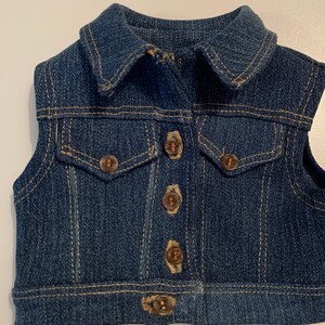 May include: A blue denim vest with a collared neckline and five brown buttons. The vest has a single pocket on the front.