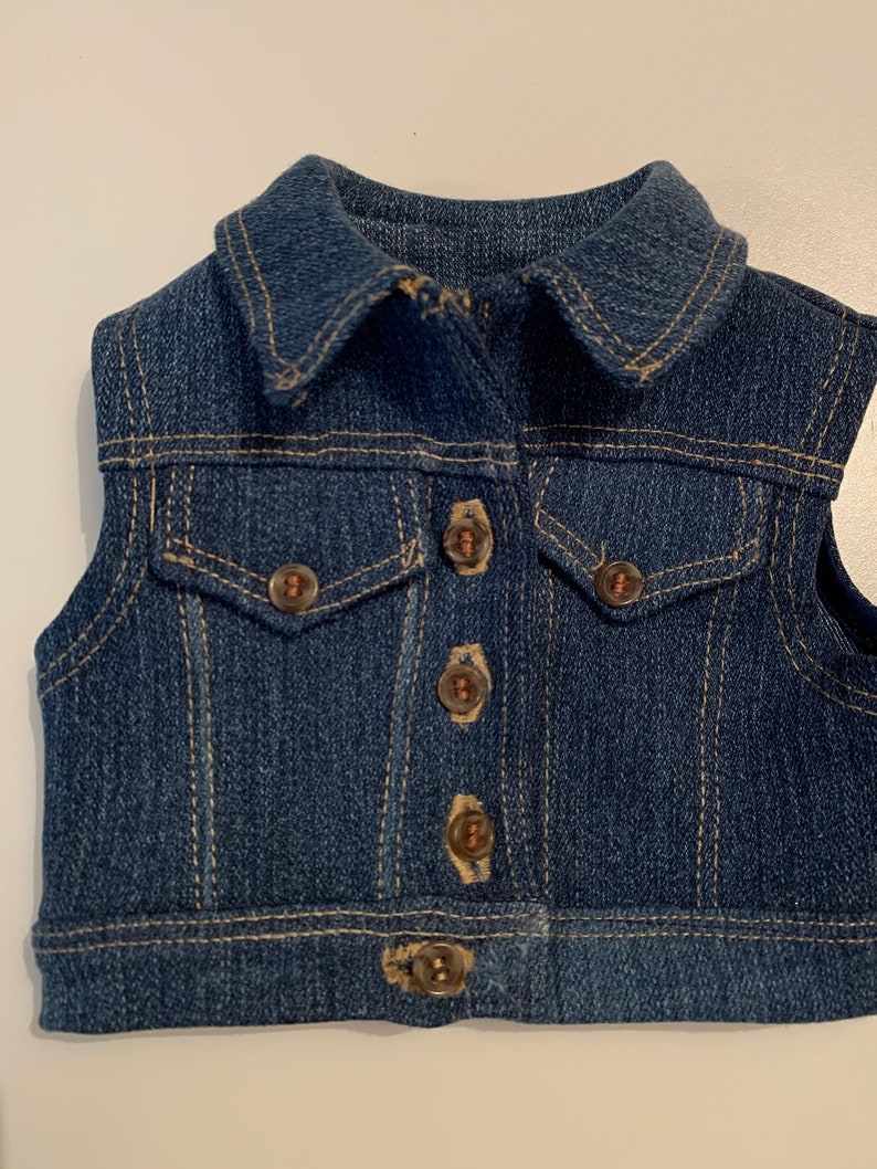 May include: A blue denim vest with a collared neckline and five brown buttons. The vest has a single pocket on the front.